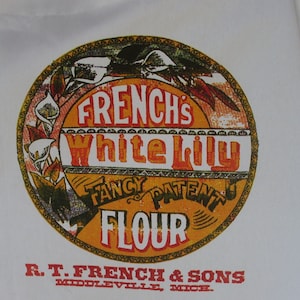 May include: White cotton apron featuring a vintage-style design. The apron has a circular emblem with the text "FRENCH'S White Lily FLOUR" in orange and red. The bottom of the apron reads "R.T. FRENCH & SONS, MIDDLEVILLE, MICH."