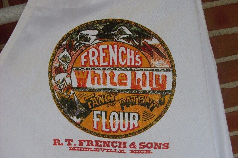 May include: White cotton apron featuring a vintage-style design. The apron has a circular emblem with the text "FRENCH'S White Lily FLOUR" in orange and red. The bottom of the apron reads "R.T. FRENCH & SONS, MIDDLEVILLE, MICH."