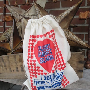 May include: A cream-colored drawstring bag with a red heart design and the words "Sweet Heart Sugar" in blue and red. The bag is decorated with small red hearts and the text "100% Pure & Natural".