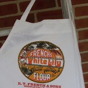 May include: White apron with a circular graphic featuring the text "FRENCH'S White Lily Fancy Patent Flour." The apron is hanging against a brick wall and a white ladder.