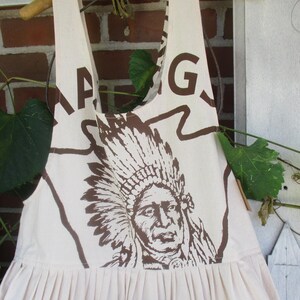 May include: A white cotton apron with a brown print of a Native American chief wearing a headdress. The apron has a gathered skirt and two straps. The text "G.A.G. 100 LB. BEST" is printed on the apron.