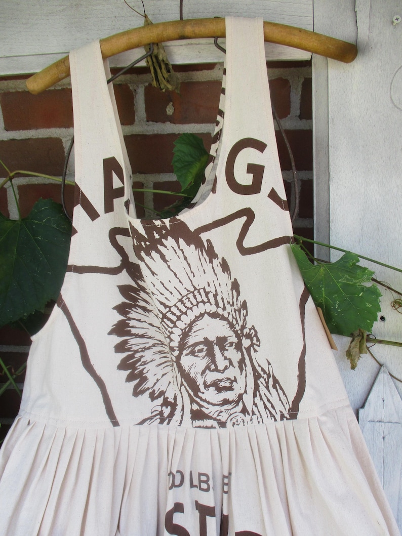 May include: A white cotton apron with a brown print of a Native American chief wearing a headdress. The apron has a gathered skirt and two straps. The text "G.A.G. 100 LB. BEST" is printed on the apron.