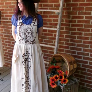 May include: A woman wearing a brown hat and a white apron with a Native American design and the text "The Farmers Poultry & Grain Co. Inc.  Distributing" printed on it.