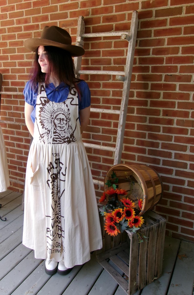 May include: A woman wearing a brown hat and a white apron with a Native American design and the text "The Farmers Poultry & Grain Co. Inc.  Distributing" printed on it.