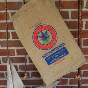 May include: A burlap sack with a blue and red label that reads "EXTRA HIGH QUALITY" and "WILD FLOWER SEED". The label also features a blue flower with a red circle around it. The sack is hanging on a rusty metal gate.