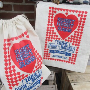 May include: Two off-white canvas bags with red heart and text graphics. One is a drawstring bag, the other a tote bag. The text reads "Sweet Heart Sugar" and "100% Pure & Natural".