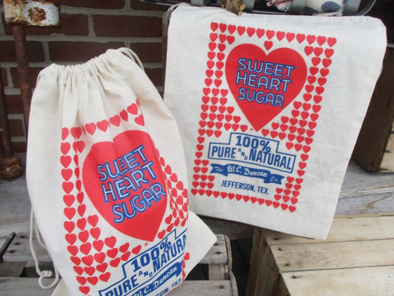 May include: Two off-white canvas bags with red heart and text graphics. One is a drawstring bag, the other a tote bag. The text reads "Sweet Heart Sugar" and "100% Pure & Natural".
