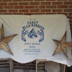 May include: A white fabric sack with blue text that reads "Fancy Blue Ribbon Race Horse Oats" and "Manufactured for H.V.R. Grain Company Pipestone, Minn. 56164". The sack is decorated with two rustic, brown, five-pointed stars.