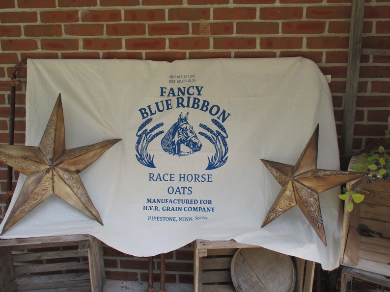 May include: A white fabric sack with blue text that reads "Fancy Blue Ribbon Race Horse Oats" and "Manufactured for H.V.R. Grain Company Pipestone, Minn. 56164". The sack is decorated with two rustic, brown, five-pointed stars.