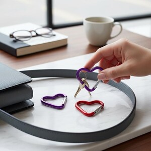 May include: Three heart-shaped carabiners in purple and red metallic finishes. One purple carabiner is held and features a silver metal key ring attached to it, while another purple carabiner and one red carabiner rest on a white surface.