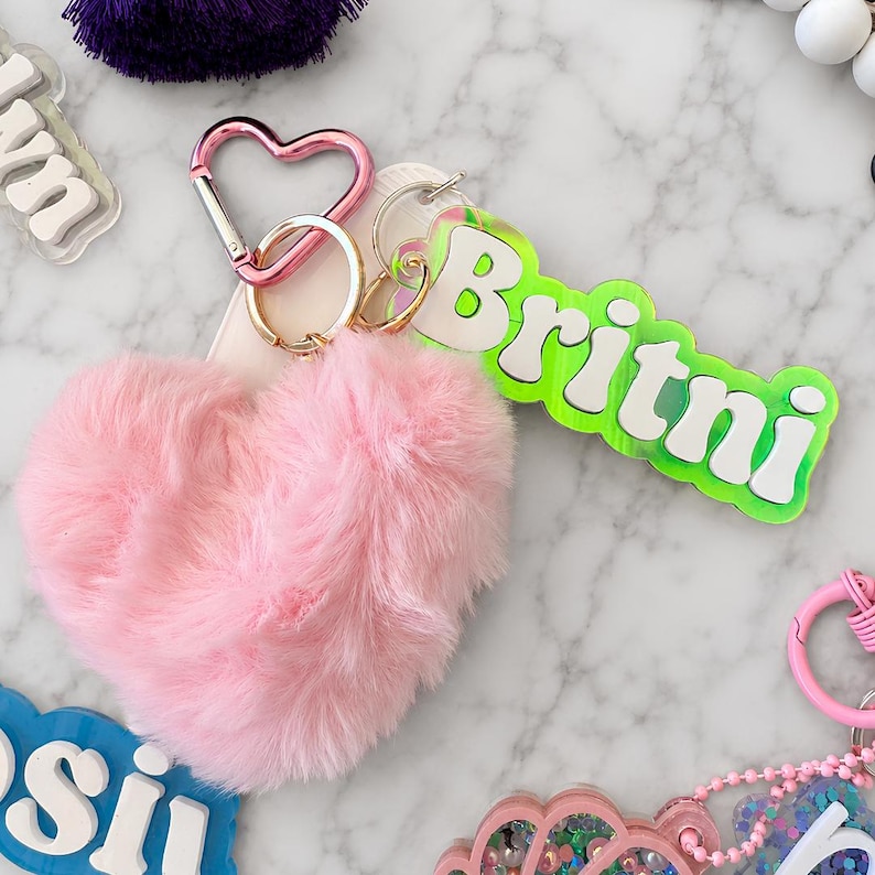 May include: A heart-shaped pink faux fur keychain attached to a pink heart-shaped carabiner and a gold-tone metal ring. A rectangular acrylic tag with a neon green border and white lettering that reads Britni is attached to the ring.