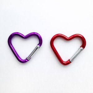 May include: Two heart-shaped carabiners, one in a vibrant purple and the other in a bright red. Each carabiner features a smooth, metallic finish and a silver-colored spring-loaded gate mechanism that allows for opening and closing the clip.