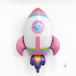 May include: A colorful rocket-shaped balloon with a white body, pink accents, and blue and yellow stripes. The balloon features a circular window with a galaxy design and pink wings. The bottom has a purple and yellow flame design.