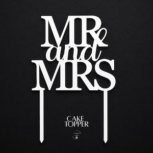 May include: White cake topper with the words "MR and MRS" in a bold, elegant font. The words are connected by the word "and" in a script font. The topper has two stakes for insertion into a cake. The words "CAKE TOPPER" are at the bottom.