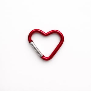 May include: A heart-shaped carabiner with a glossy red finish and a silver-toned spring-loaded gate mechanism. The metal clip features a smooth, rounded frame that forms a distinct heart outline, providing a functional and secure attachment point.
