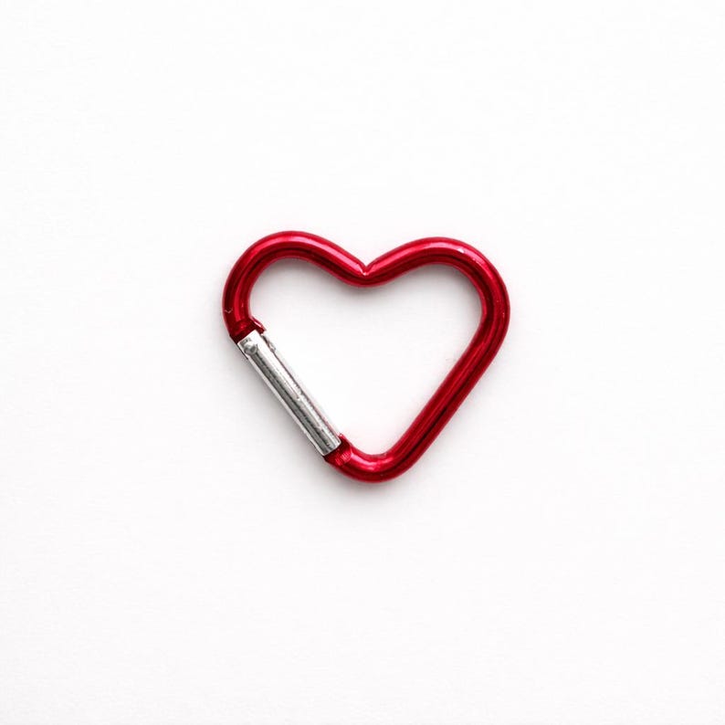 May include: A heart-shaped carabiner with a glossy red finish and a silver-toned spring-loaded gate mechanism. The metal clip features a smooth, rounded frame that forms a distinct heart outline, providing a functional and secure attachment point.
