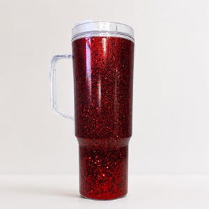 May include: A tall, cylindrical tumbler with a clear plastic handle and a matching clear lid. The exterior of the cup is covered in a dense, vibrant red glitter finish that creates a textured, sparkling appearance across the entire surface.