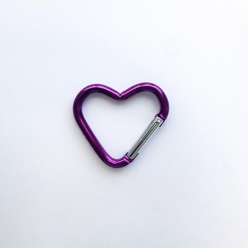 May include: A heart-shaped carabiner with a metallic purple finish and a silver-toned spring-loaded gate mechanism. The carabiner features a smooth, rounded frame that forms a distinct heart shape, providing a functional clip for various attachments.