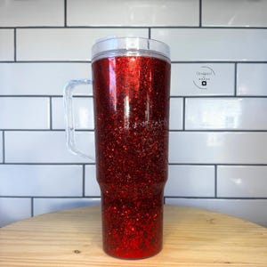 May include: A tall, red glitter tumbler with a clear handle and lid. The tumbler is filled with sparkling red glitter and has a clear handle for easy carrying. The tumbler is designed to keep drinks cold or hot.