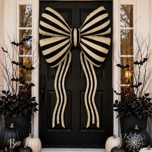 May include: A large black and cream striped fabric bow attached to a black door. The door is flanked by two black planters containing bare branches with black bat cutouts. Several black and white pumpkins are arranged on the ground.