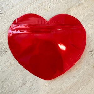 May include: A bright red, heart-shaped pouch with a sealed top. The pouch is made of a shiny, reflective material. The background is a light-colored wooden surface.