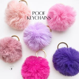 May include: A collection of fluffy, colorful keychains in shades of pink, purple, and fuchsia. Each keychain features a round, furry pom-pom with a gold-colored metal ring. The text "POOF KEYCHAINS" is displayed in the upper center of the image.