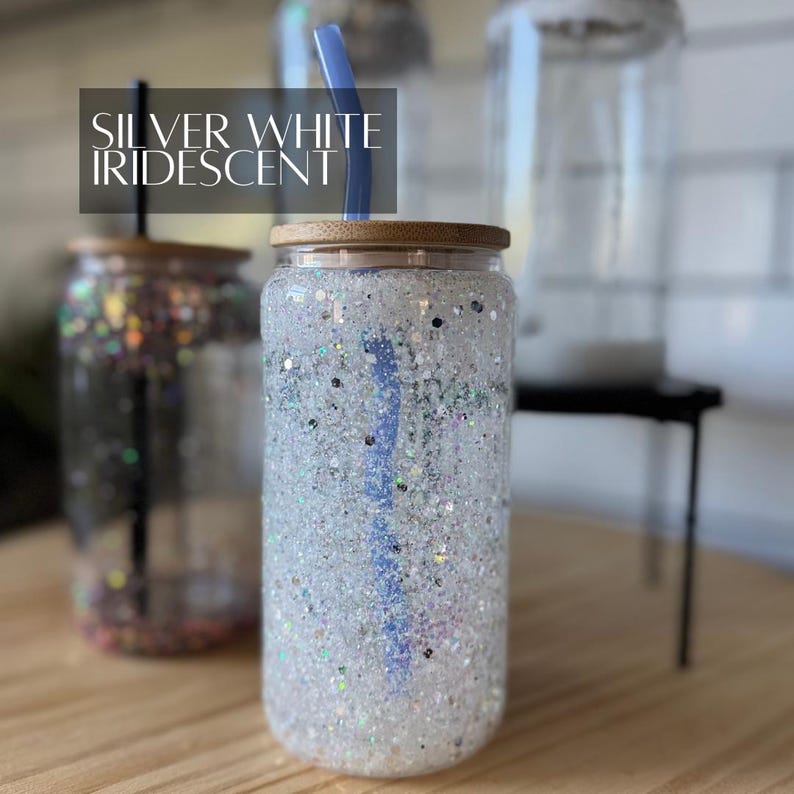 May include: A clear glass tumbler with a silver and white iridescent glitter design. The tumbler has a bamboo lid and a blue straw. The text "SILVER WHITE IRIDESCENT" is visible in the image.