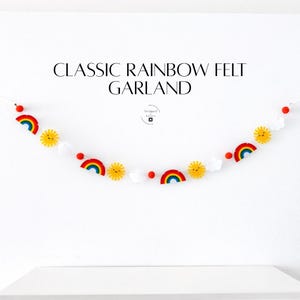 May include: A felt garland featuring a repeating pattern of rainbows, suns, and clouds. The garland is strung on a white string and includes the text "CLASSIC RAINBOW FELT GARLAND". The colors include red, yellow, blue, and white.