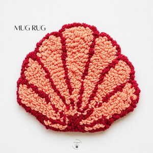 May include: A handmade mug rug in the shape of a seashell. The rug is made of a textured, looped yarn in shades of coral and burgundy. The text "MUG RUG" is printed in black at the top of the image.