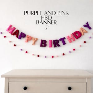 May include: A purple and pink "Happy Birthday" banner with felt letters and pom-poms. The banner is strung across a white wall above a light-colored dresser. The text "Purple and Pink HBD Banner" is above the banner.