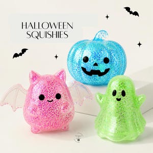 May include: Three Halloween squishies featuring a blue glittery jack-o'-lantern, a pink glittery bat with outstretched wings, and a green glittery ghost. Each figure has black eyes and a simple smile. The text HALLOWEEN SQUISHIES is visible.