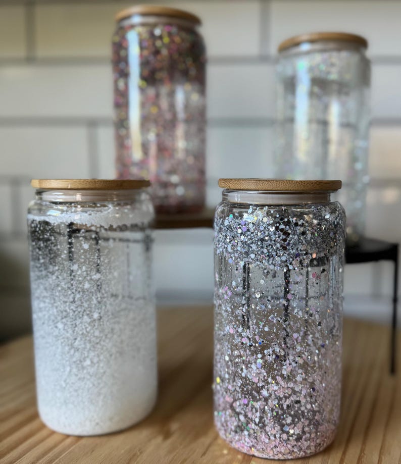 May include: Four glass jars with wooden lids filled with glitter. The jars are clear and the glitter is a variety of colors, including silver, gold, and pink.
