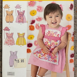 May include: Simplicity sewing pattern 1470A for baby dresses and rompers. The pattern includes multiple dress and romper designs with matching hats. The image shows a baby wearing a pink gingham dress with a floral apron.