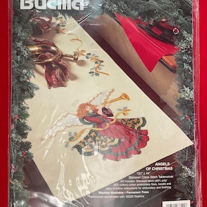 May include: A Bucilla Angels of Christmas cross stitch tablerunner kit. The kit includes stamped fabric, embroidery floss, a needle, and instructions. The design features an angel playing a trumpet with a red, green, and gold color scheme. The tablerunner measures 13 inches by 44 inches.