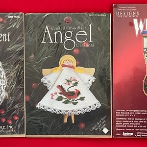 May include: Three cross-stitch ornament kits. The first is an oval lace ornament with a teddy bear and the word "NOEL". The second is an angel ornament. The third is a stocking ornament with a teddy bear and the name "John".