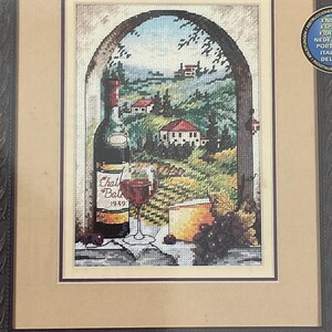 May include: A framed cross-stitch kit titled Dreaming of Tuscany, measuring 5 inches by 7 inches. The design features a wine bottle, a glass of red wine, cheese, and grapes set against an arched window view of a rolling landscape with houses.