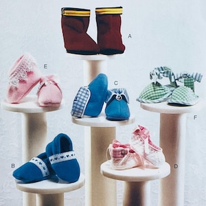 May include: A collection of baby shoes in various styles and colors. The selection includes red boots, blue moccasins, pink booties with lace, and green plaid sandals. Each pair is displayed on a small, white pedestal.