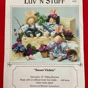 May include: A printed page featuring three seated bunny dolls with floral print clothing and bunny ears. The dolls are on a wooden crate with flowers and carrots. The text reads "Luv 'N Stuff" and "Sweet Violets". The dolls are approximately 12 inches tall.