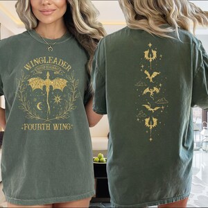 A woman wearing a green t-shirt with a dragon design and text on it, standing in a room with a window and a couch visible in the background.