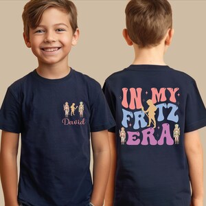 May include: A navy blue t-shirt featuring a small graphic on the front left chest with three nutcracker figures and the name David in pink script. The back displays the text IN MY FRITZ ERA in large, wavy, multi-colored letters with nutcrackers.