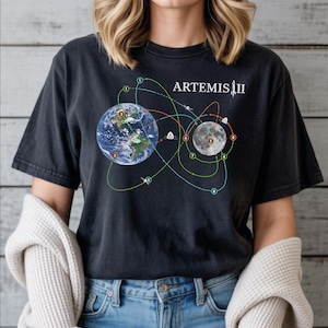 May include: Black t-shirt featuring a graphic of Earth and the Moon connected by orbital paths, with the text "ARTEMIS III" in white. The design includes numbered points and small spacecraft illustrations. The shirt is made of a dark, possibly faded, fabric.