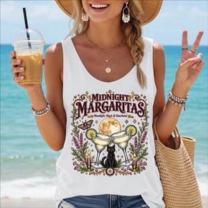 May include: A white sleeveless tank top featuring a graphic print with the text "MIDNIGHT MARGARITAS" and "Moonlight, Magic & Sisterhood". The design includes two margarita glasses, a black cat, a full moon, a moth, candles, and floral elements.
