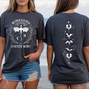 A woman wearing a black t-shirt with a graphic design featuring a bat and stars on the front and back, standing on a beach with the ocean in the background.