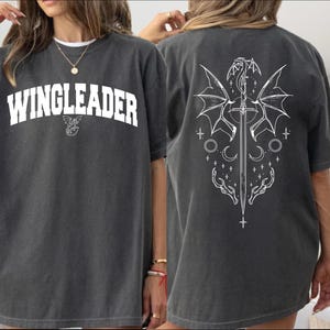 A woman wearing a grey t-shirt with the text "WINGLEADER" printed on it, along with a graphic design featuring a winged creature and a sword.