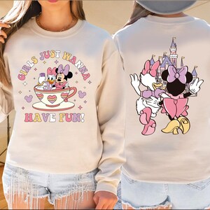 A woman wearing a white sweatshirt with a cartoon image of Minnie Mouse and Donald Duck on it, along with the text "Girls just wanna have fun!" printed on it.
