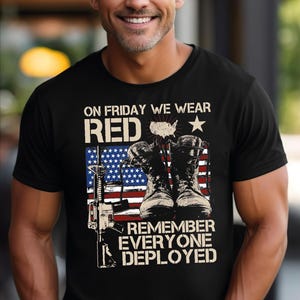 May include: Black t-shirt with the text "ON FRIDAY WE WEAR RED" above an American flag design, combat boots, and a rifle. The text "REMEMBER EVERYONE DEPLOYED" is below the flag. The shirt is worn by a person.