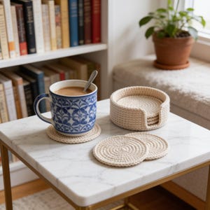 May include: A set of round, cream-colored crocheted coasters with a spiral pattern. One coaster sits under a white and blue patterned ceramic mug containing a brown beverage with a metal spoon, while others are stacked in a matching holder.