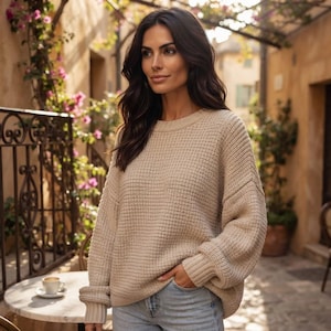 Könnte beinhalten: Ein beigefarbener Langarmpullover mit grober Strickstruktur, Rundhalsausschnitt und gerippten Bündchen. Der Pullover hat eine lockere Passform mit überschnittenen Schultern und wird mit einer hellblauen Jeans kombiniert für einen neutralen Look.