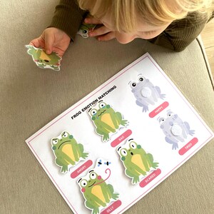 K&ouml;nnte beinhalten: Eine wei&szlig;e Karte mit den Worten "Frog Emotion Matching" darauf gedruckt. Es gibt sechs gr&uuml;ne Fr&ouml;sche mit verschiedenen Gesichtsausdr&uuml;cken, die jeweils mit einer Emotion beschriftet sind: w&uuml;tend, traurig, &auml;ngstlich, albern, gl&uuml;cklich und m&uuml;de.