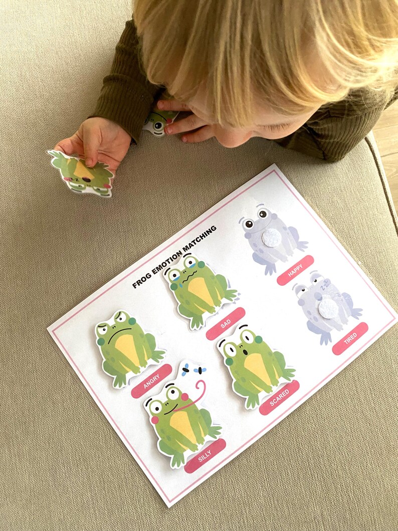 K&ouml;nnte beinhalten: Eine wei&szlig;e Karte mit den Worten "Frog Emotion Matching" darauf gedruckt. Es gibt sechs gr&uuml;ne Fr&ouml;sche mit verschiedenen Gesichtsausdr&uuml;cken, die jeweils mit einer Emotion beschriftet sind: w&uuml;tend, traurig, &auml;ngstlich, albern, gl&uuml;cklich und m&uuml;de.